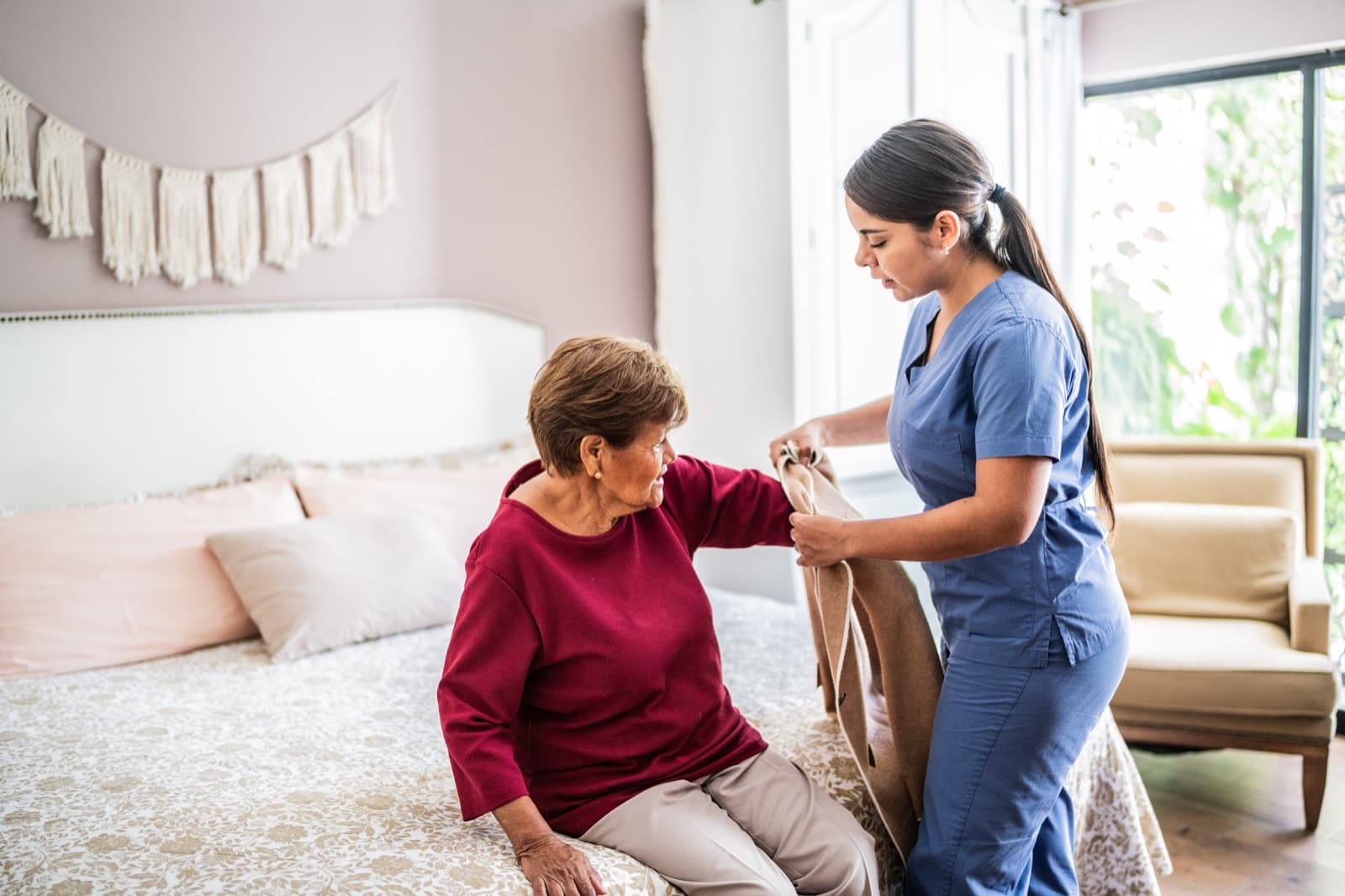 Caregiver helping an older adult get dressed in a bright bedroom at home
