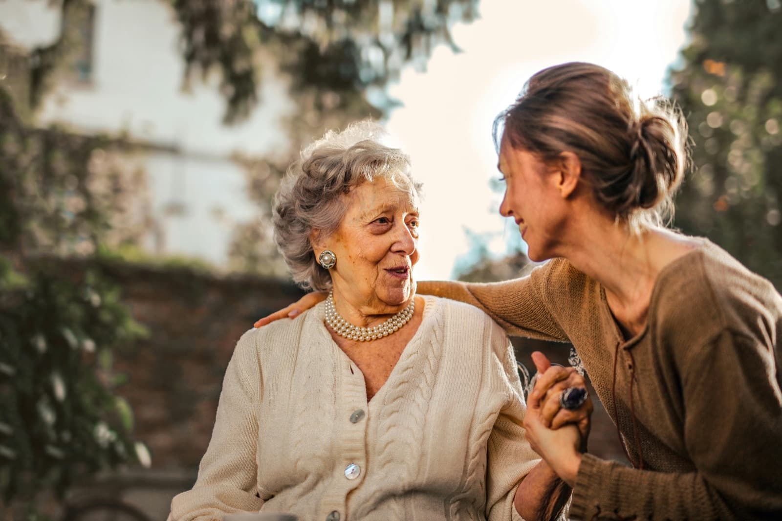 Elderly woman and younger companion smiling warmly while holding hands in a sunlit garden