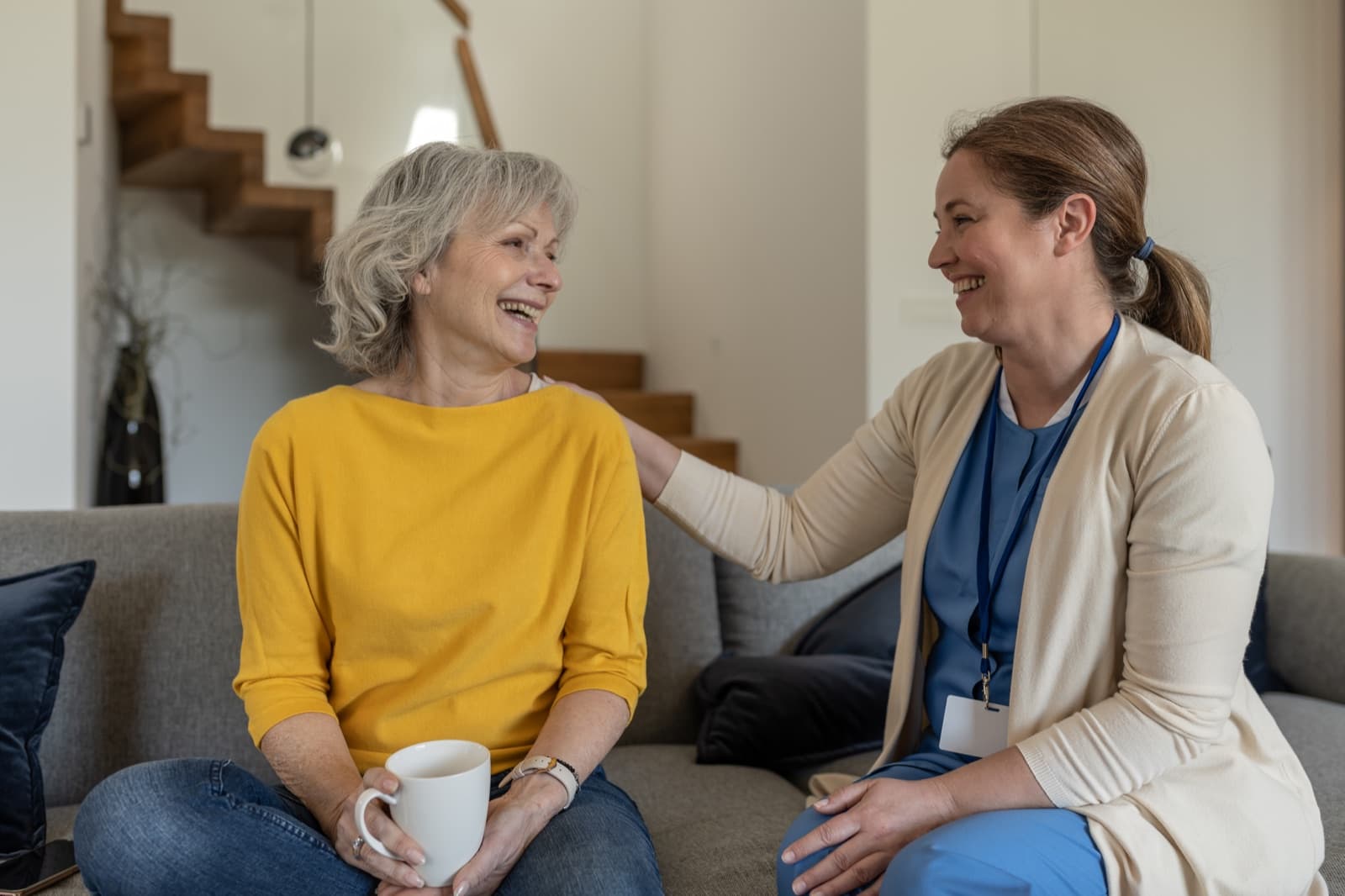 Caregiver and older woman sharing a warm conversation over coffee on a comfortable sofa in a bright home
