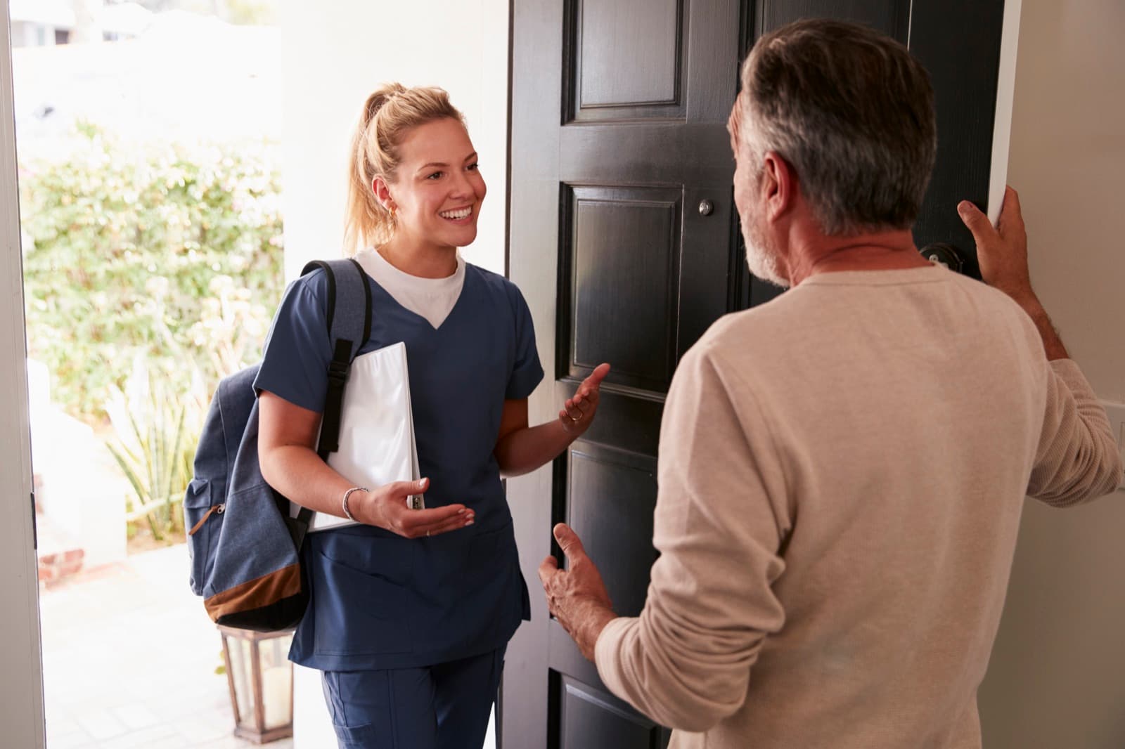 Friendly visitor with a backpack and notebook smiling as an older man opens his front door to welcome her