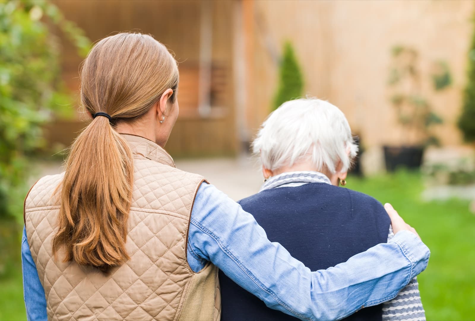 Caregiver walking beside an older adult with a supportive hand on her shoulder outdoors